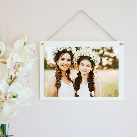 A white coated wooden rectangular hanging sign from the PhotoSplash HomeSplash collection, suspended by rustic twine and showcasing a photo, logo, or monogram with outstanding color reproduction on the wall with flowers.